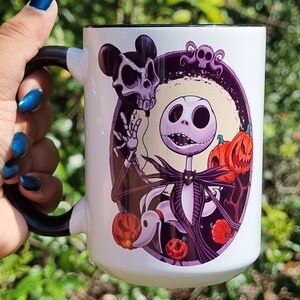 May include: White ceramic mug with a black handle and rim, featuring a colorful illustration of Jack Skellington, Zero, pumpkins, and a skull. The design is set against a background of a purple oval. A spooky themed mug.