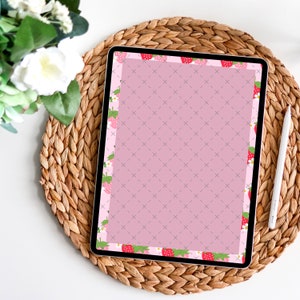 Könnte beinhalten: Ein digitales Tablet mit rosa Bildschirm und Erdbeer-Randdesign. Das Tablet liegt auf einem gewebten Platzset mit einem weißen Stift und einem weißen Blumenarrangement. Der Erdbeer-Rand umfasst rote Erdbeeren und grüne Blätter.