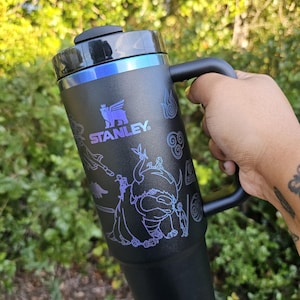 May include: Black Stanley travel mug with a handle, featuring a purple graphic design of characters and symbols. The mug has a black lid and a blue accent around the top. The Stanley logo is visible.
