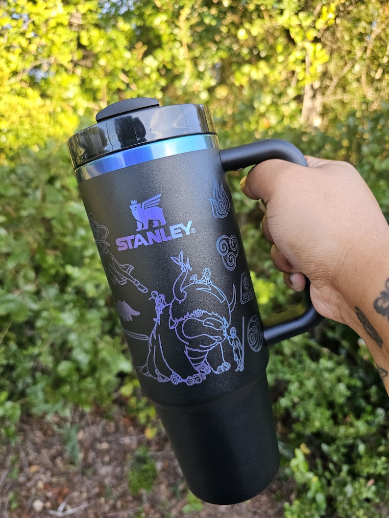 May include: Black Stanley travel mug with a handle, featuring a purple graphic design of characters and symbols. The mug has a black lid and a blue accent around the top. The Stanley logo is visible.