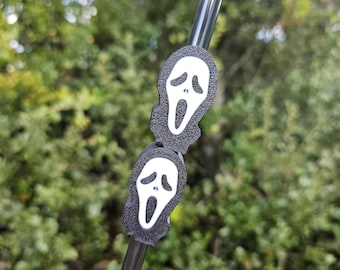 May include: A black and clear reusable straw with two Scream mask charms. The charms are black with a white ghost face design. The background is blurred green foliage.
