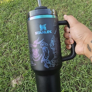 Puede incluir: Vaso Stanley negro con asa y pajita, con una banda turquesa y un diseño de Harry Potter. El diseño incluye ilustraciones de Hogwarts, un escudo y otros elementos mágicos. El logotipo de Stanley es visible.