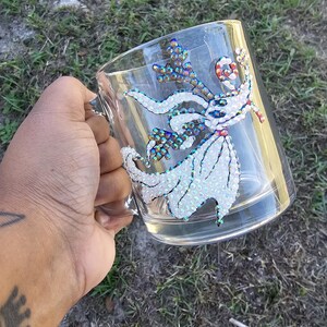 May include: Clear glass mug decorated with a sparkling Zero character design. The design features white, iridescent, and colorful rhinestones. The mug has a handle and is held against a background of green grass.