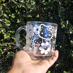 May include: Clear glass mug featuring white floral and Hello Kitty designs. The mug has a handle and is held in a hand against a backdrop of green foliage. The design includes multiple white flowers and the iconic Hello Kitty face.