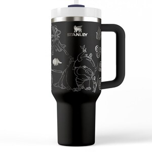May include: Black Stanley tumbler with a white lid, handle, and straw. The tumbler features a detailed white graphic design of various characters and the Stanley logo. The tumbler is designed to keep beverages cold or hot.