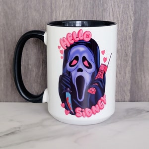May include: White ceramic mug with a black handle and interior. The mug features a graphic of a ghost-faced figure holding a knife and a retro phone, with the words "Hello Sidney" in pink.
