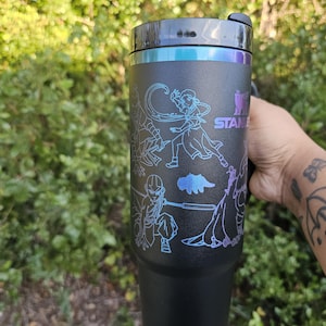May include: Black Stanley tumbler with a black lid and a silver rim. The tumbler features a design of characters in a blue and purple gradient. The Stanley logo is visible. The tumbler is held by a person.