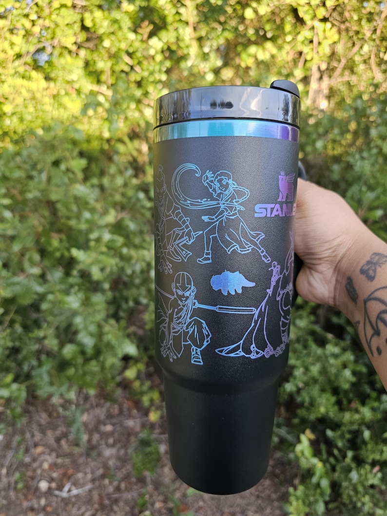 May include: Black Stanley tumbler with a black lid and a silver rim. The tumbler features a design of characters in a blue and purple gradient. The Stanley logo is visible. The tumbler is held by a person.