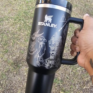 May include: Black Stanley travel mug with a handle, featuring etched designs of a house, a creature, and other graphics. The mug has a black lid and a silver rim. The Stanley logo is visible. The mug is held by a person.