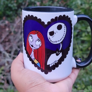 May include: White ceramic mug featuring a heart-shaped design with illustrations of Sally and Jack Skellington from The Nightmare Before Christmas. The mug has a black handle and rim, and is held in a hand.