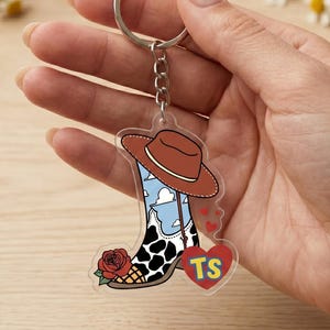 May include: A clear acrylic keychain featuring a cowboy boot design with a brown hat, a blue sky with white clouds, a black and white cow print pattern, a red rose, and a red heart shape containing the yellow letters TS.
