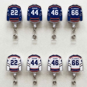 May include: Eight hockey jersey-shaped retractable badge holders. The jerseys are white, blue, and red, with player names and numbers. The names include Logan, Graham, Tucker, and Di Laurentis. Each badge holder has a clear plastic strap and a metal clip.
