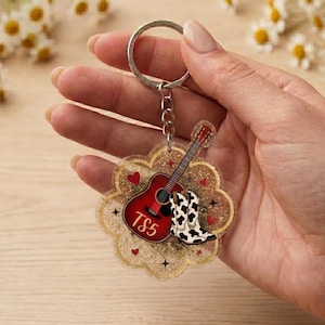 May include: A clear acrylic keychain with a scalloped edge filled with gold glitter. The design features a red acoustic guitar with the text TS5, a pair of black and white cow print cowboy boots, and small red hearts and black star accents.