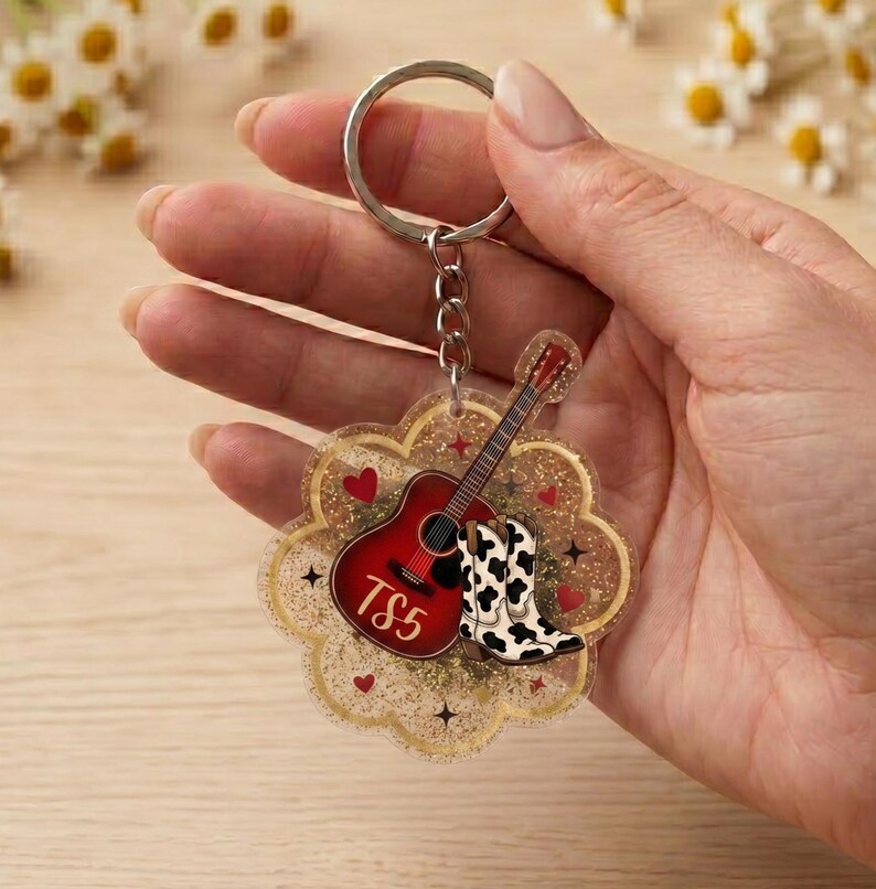 May include: A clear acrylic keychain with a scalloped edge filled with gold glitter. The design features a red acoustic guitar with the text TS5, a pair of black and white cow print cowboy boots, and small red hearts and black star accents.