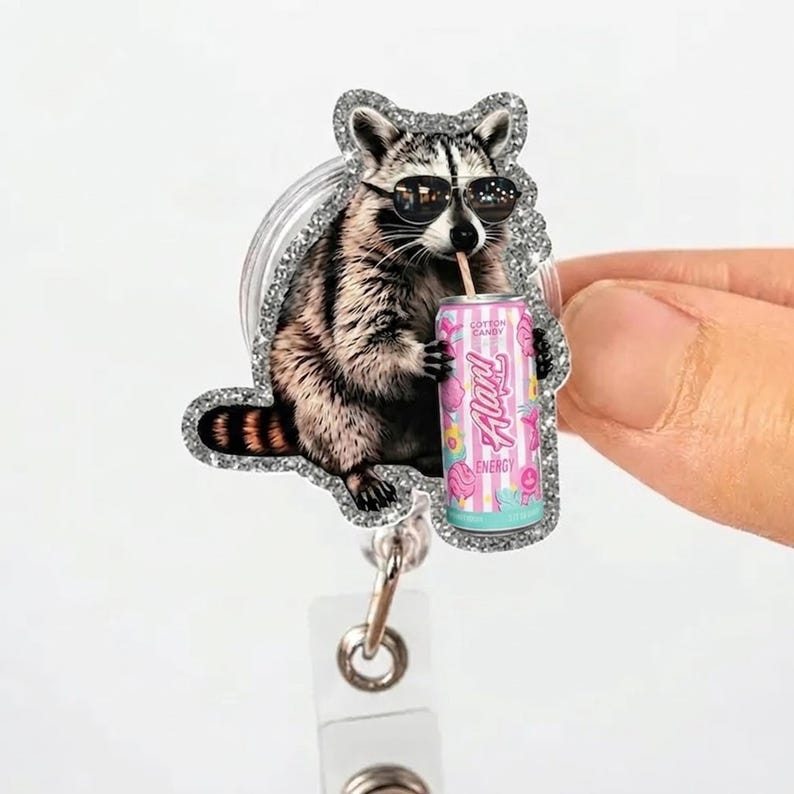 May include: A badge reel featuring a raccoon wearing sunglasses and drinking from a pink and blue can labeled "Alani Energy." The raccoon is surrounded by a glittery silver border. The can has the words "Cotton Candy" on it.