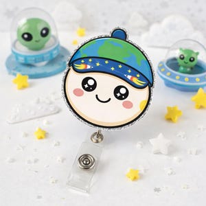 Puede incluir: Un portatarjetas redondo de estilo caricatura con una cara sonriente que lleva un sombrero con el tema de la Tierra. El portatarjetas tiene un borde plateado brillante y un clip de plástico transparente. Figuras de extraterrestres y decoraciones en forma de estrella en el fondo.