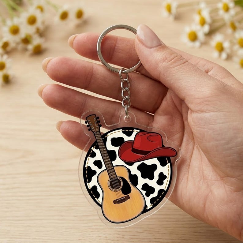 May include: A circular acrylic keychain featuring a brown acoustic guitar and a red cowboy hat set against a black and white cow print background. The design is attached to a silver metal split ring by a short chain, creating a decorative accessory.