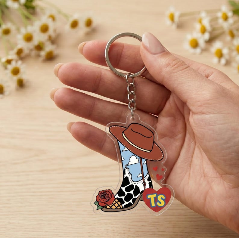 May include: A clear acrylic keychain featuring a cowboy boot design with a brown hat, a blue sky with white clouds, a black and white cow print pattern, a red rose, and a red heart shape containing the yellow letters TS.