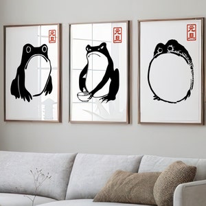 May include: Three framed art prints featuring black frog illustrations on white backgrounds. Each print has a different frog pose, with a red Japanese character in the upper right corner. The frames are a warm brown color.