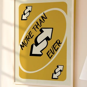 May include: Framed art print featuring a yellow card design with the words "MORE THAN EVER" and white arrows. The artwork is displayed on a wall, with a lamp and decorative items on a table below.