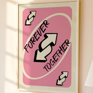 May include: Framed art print featuring a pink card design with the words "FOREVER TOGETHER" and white arrows. The artwork is in a white frame and hangs on a light-colored wall. A lamp and decorative items are visible on a table below.
