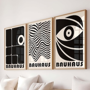 May include: Three framed Bauhaus art prints with black and white geometric designs. The prints are displayed above a wooden bench with white cushions and a woven basket. Each print has the word "BAUHAUS" at the bottom.