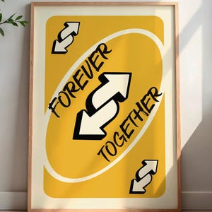 May include: Framed art print featuring a yellow card design with the words "FOREVER TOGETHER" and white arrows. The artwork is in a wooden frame and is displayed in a room with a side table and a vase with greenery.