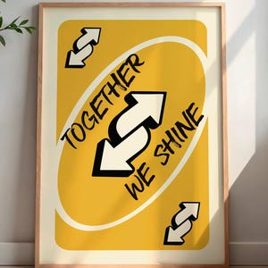 May include: Framed art print featuring a yellow card design with the text "TOGETHER WE SHINE" and reverse arrows. The artwork is in a wooden frame and is displayed indoors, leaning against a wall. A decorative vase and side table are visible.