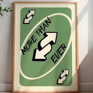 May include: Framed art print featuring a green card design with the words "MORE THAN EVER" and white arrows. The artwork is in a wooden frame and is displayed against a neutral wall. A decorative vase and side table are visible in the background.