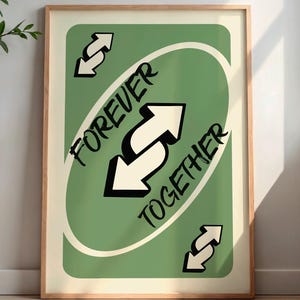 May include: A framed art print featuring a green card design with the words "FOREVER TOGETHER" in black text. The card has white arrows and a cream-colored background. The frame is a light wood color.