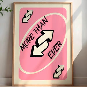 May include: Framed art print featuring a pink card design with the words "MORE THAN EVER" and reverse arrows. The artwork has a light beige border and is displayed in a wooden frame. The print is set against a neutral wall.