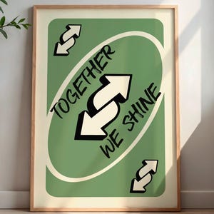 May include: A framed poster featuring a green card design with the text "TOGETHER WE SHINE" and reverse arrows. The poster is in a wooden frame and is displayed against a neutral wall. A small wooden side table with a white vase and plant is to the left.