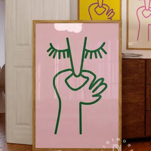 May include: A pink poster with a green line drawing of a face with closed eyes and a hand covering the mouth. The poster is framed in wood and is hanging on a wall. The text 'DIGINORM DESIGN' is printed in white on a black background at the bottom of the poster.