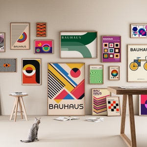 May include: A collection of framed Bauhaus-style posters with geometric shapes, bold colors, and the word "Bauhaus" in various fonts. The posters are displayed on a white wall with a wooden table and stool in the foreground.