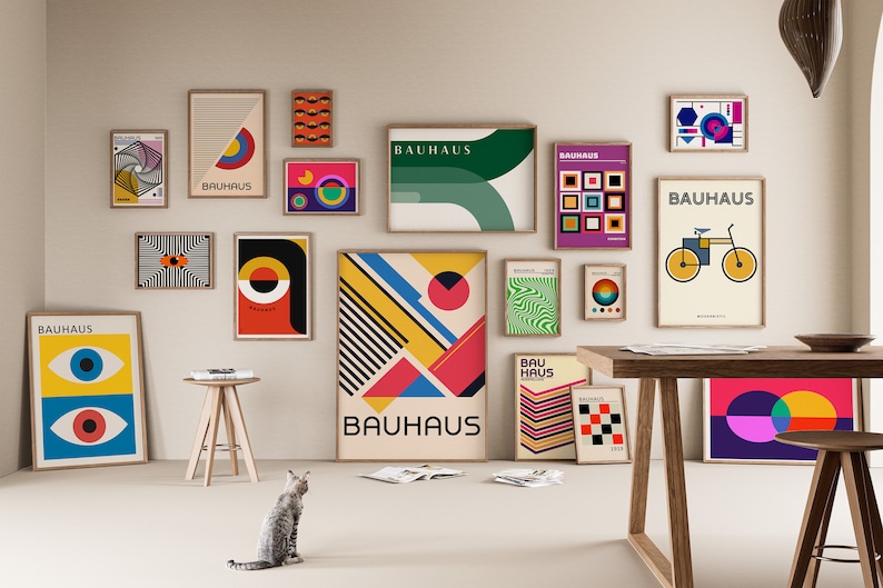 May include: A collection of framed Bauhaus-style posters with geometric shapes, bold colors, and the word "Bauhaus" in various fonts. The posters are displayed on a white wall with a wooden table and stool in the foreground.