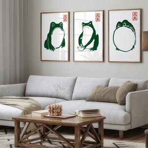 May include: Three framed prints with green frog illustrations. The frogs are in different poses, one is holding a bowl, one is sitting with its legs crossed, and one is in a circle shape. The frames are brown wood and the background is a light gray wall.