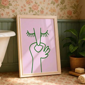 May include: A framed art print featuring a minimalist line drawing of a face with closed eyes and a hand. The artwork is rendered in green on a pink background, set within a light wood frame. The print is displayed in a bathroom setting.