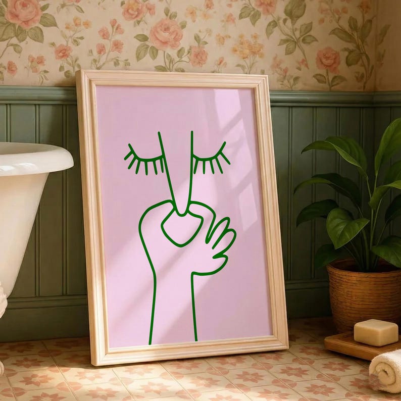 May include: A framed art print featuring a minimalist line drawing of a face with closed eyes and a hand. The artwork is rendered in green on a pink background, set within a light wood frame. The print is displayed in a bathroom setting.
