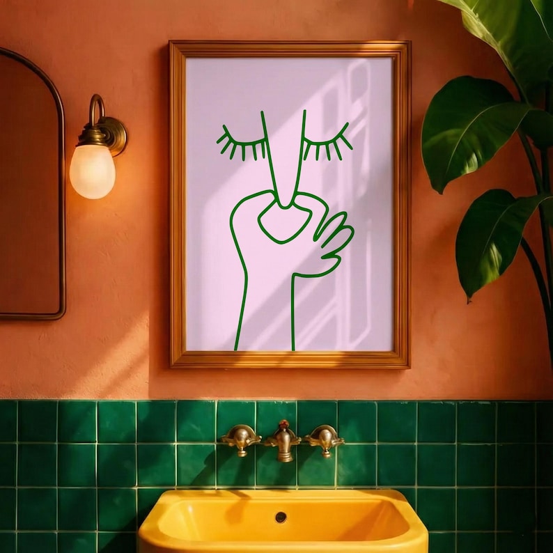 May include: A framed art print featuring a minimalist line drawing of a face with closed eyes and a hand. The artwork is in green on a pink background, set in a gold frame. The bathroom decor includes a yellow sink and green tile.