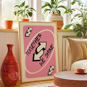 May include: A framed pink print with the words "TOGETHER WE SHINE" and two black and white arrows. The print is in a light wood frame. A red vase and a small orange cup and saucer are in the foreground.