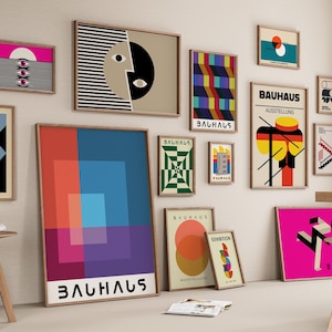 Puede incluir: Una pared de galería con varias impresiones enmarcadas que presentan formas geométricas, diseños abstractos y la palabra "Bauhaus" en diferentes colores y fuentes. Las impresiones están dispuestas en un patrón de cuadrícula en una pared blanca.
