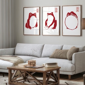 Puede incluir: Tres impresiones enmarcadas con ilustraciones de ranas rojas. Las impresiones están colgadas en una pared blanca sobre un sofá seccional gris claro con un cojín de tiro marrón. Las impresiones presentan los caracteres chinos para "buena suerte".