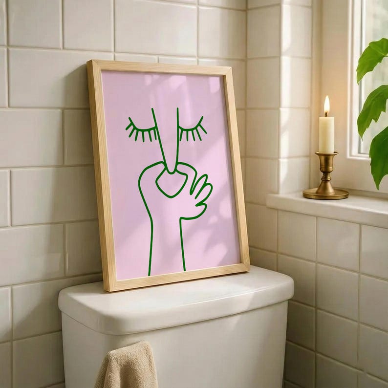 May include: A framed art print featuring a minimalist line drawing of a face with closed eyes and a hand gesture. The artwork is in green on a pink background, set within a light wood frame. The print is displayed on a white toilet.