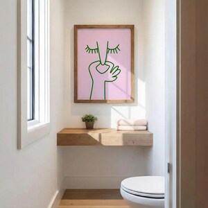 May include: A framed art print featuring a green line drawing of a face and hand on a pink background. The artwork is displayed in a wooden frame. The print is hanging above a wooden shelf in a bathroom setting.