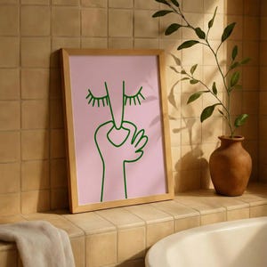 May include: A framed art print featuring a minimalist line drawing of a face with closed eyes and a hand. The artwork is in green on a pink background, set within a light wood frame. The print is displayed in a bathroom setting.
