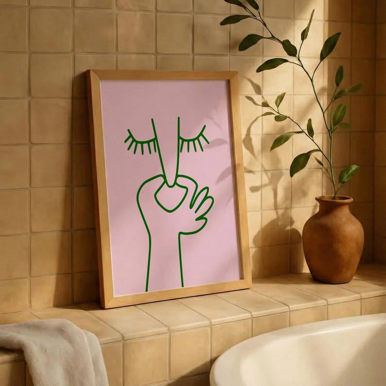 May include: A framed art print featuring a minimalist line drawing of a face with closed eyes and a hand. The artwork is in green on a pink background, set within a light wood frame. The print is displayed in a bathroom setting.
