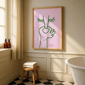 May include: A framed art print featuring a minimalist line drawing of a face and hand in green on a pink background. The artwork is displayed in a bathroom setting, with a wooden stool, towels, and a black and white checkered floor.
