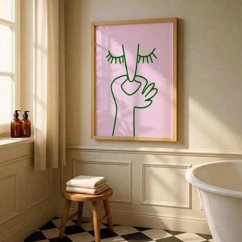 May include: A framed art print featuring a minimalist line drawing of a face and hand in green on a pink background. The artwork is displayed in a bathroom setting, with a wooden stool, towels, and a black and white checkered floor.