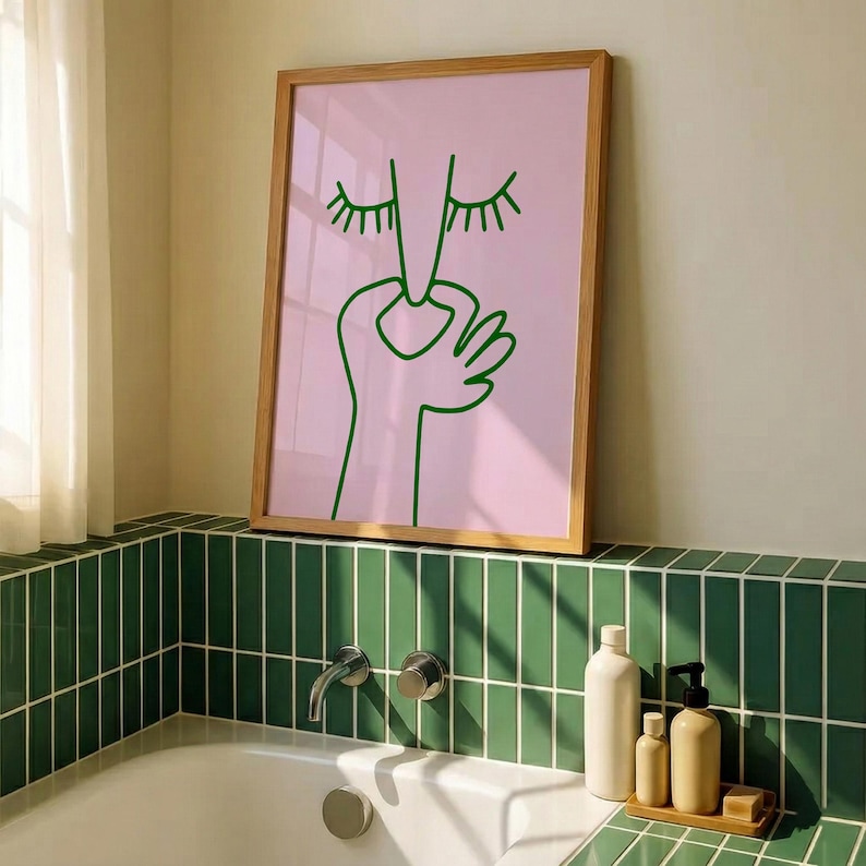 May include: A framed art print featuring a minimalist line drawing of a face and hand in green on a pink background. The artwork is in a light brown wooden frame, displayed above a green tiled surface.