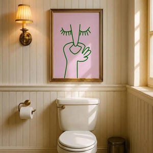 May include: A framed art print featuring a minimalist line drawing of a face with closed eyes and a hand, in green on a pink background. The artwork is in a gold frame, hanging above a white toilet in a bathroom.